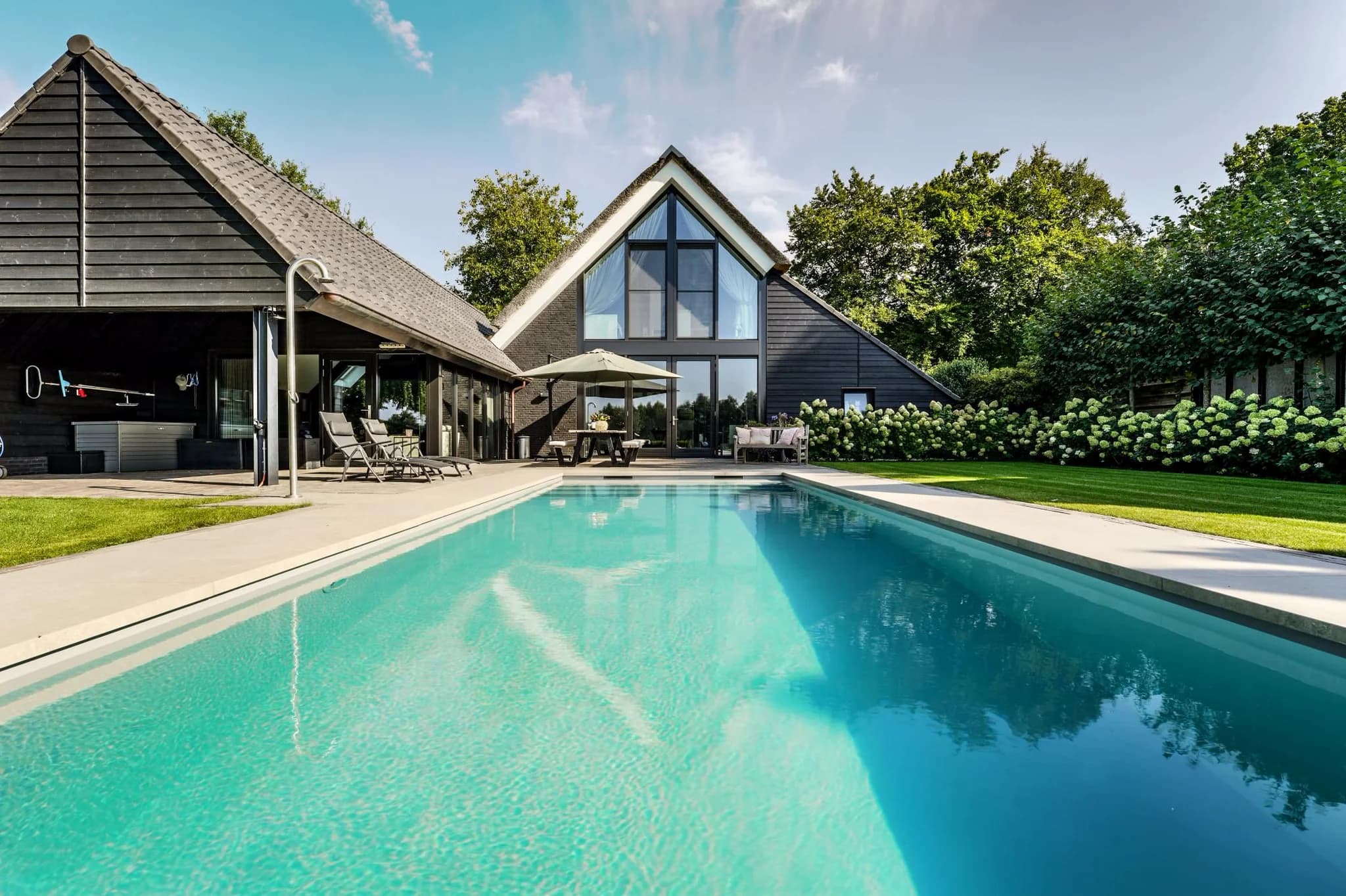 Moderne woning met glazen gevel, terras, ligstoelen en groot buitenzwembad in een groene tuin