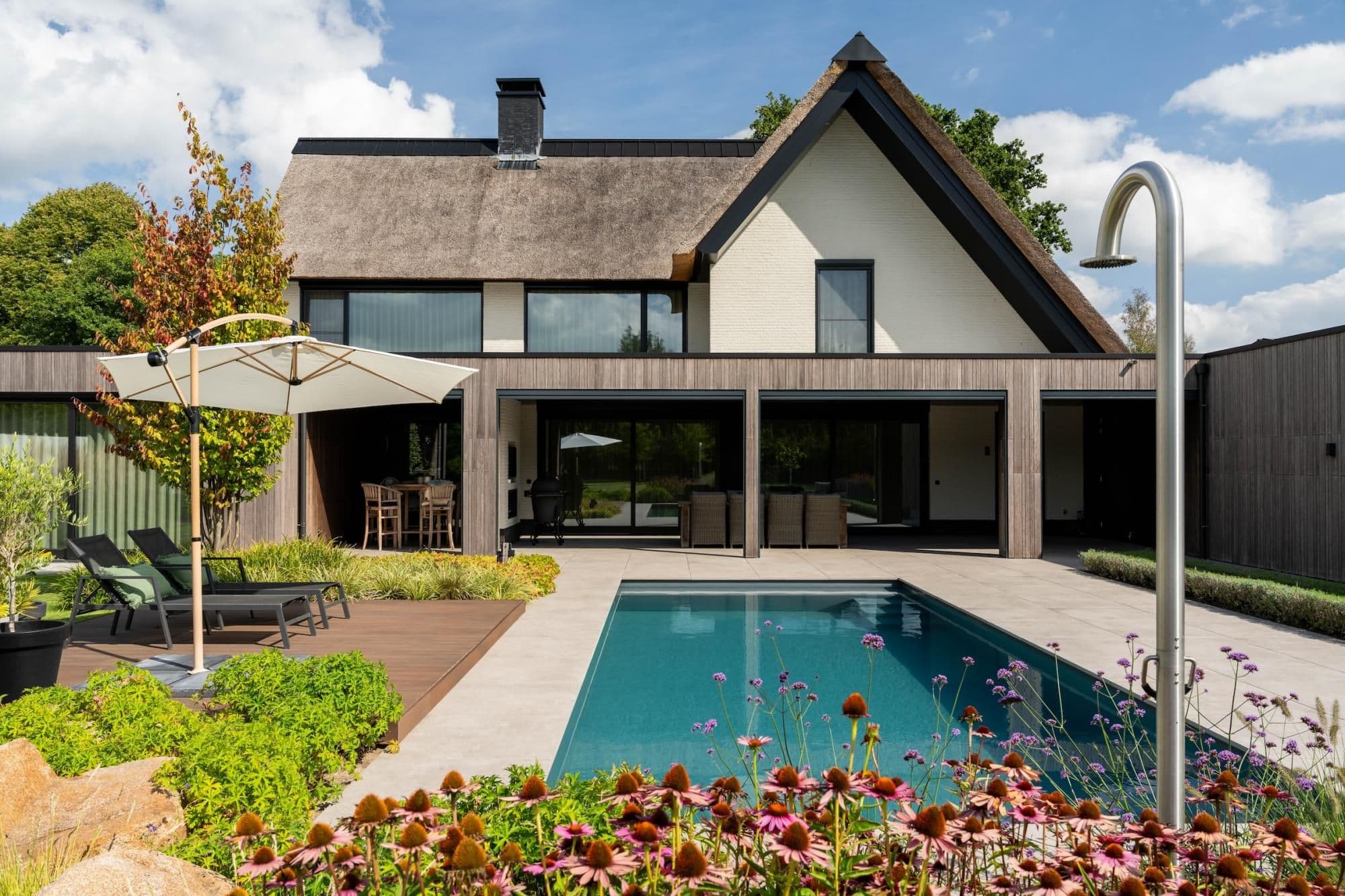 Voortuin en terras van een huis met rieten dak en houten gevel, ligstoelen en parasol naast een zwembad; buitendouche rechts in beeld.